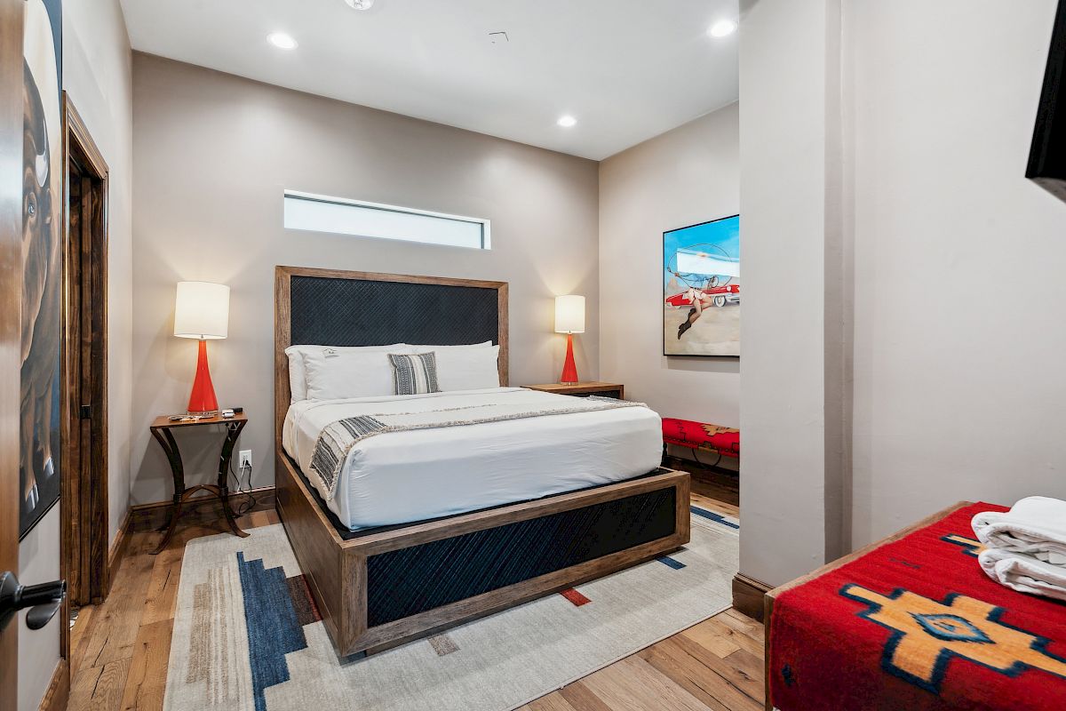 A stylish hotel room with a king bed, two lamps on side tables, a red bench at the end, and a colorful rug underfoot.