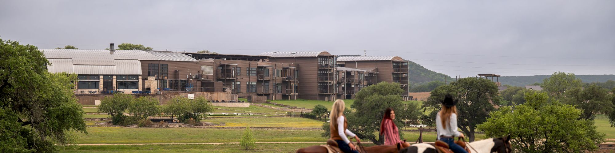 Three people ride horses along a grassy field near water, with a modern building complex in the background on a cloudy day.