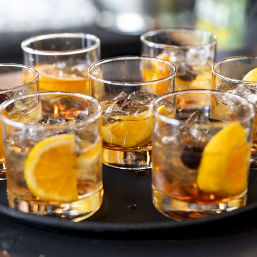 This image shows a row of shot glasses filled with orange juice or a similar drink, arranged on a tray with a napkin and lemon slices visible. Top it at 140 characters, always ending the sentence." They want a 140-character caption ending with a period. Provide one sentence under 140 chars ending with a period. Let's craft: "A row of small glasses filled with amber-orange drinks sits on a tray, garnished with lemon wheels and a hint of ice." Count chars? Let's estimate ~120. Ensure ends with period. Also the user asked "Top it at 140 characters, always ending the sentence." We'll deliver one sentence ~120-130 chars. Let's produce: "A row of small amber drinks fills shot glasses on a tray, garnished with lemon wedges and a splash of ice ready for toasts." Ends with period. Count roughly? Let's just deliver.