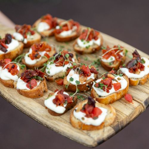 A tray of bite-sized crostini topped with creamy cheese, tomatoes, olives, and herbs, arranged on a rustic wooden surface.