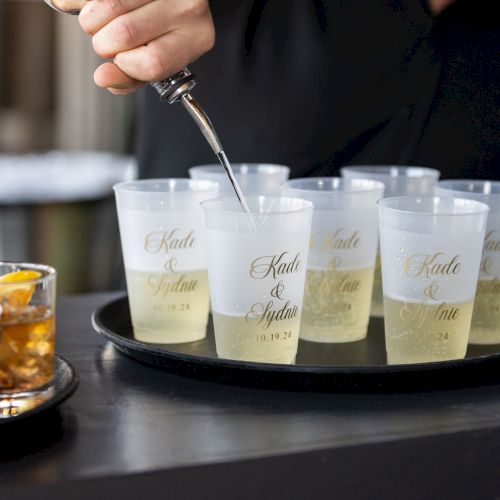 A person&rsquo;s hand pours a drink or shot into small clear cups lined up on a bar counter, with a glass of whiskey nearby.