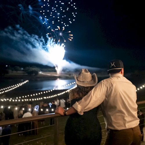 Two people watch a fireworks show from a waterfront viewing area at night, with lights and a bridge in the distance.