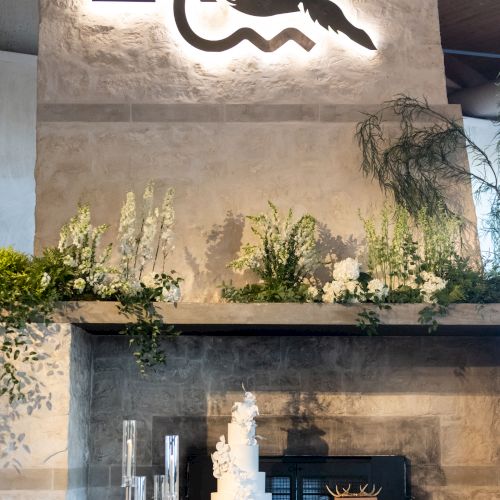 A stylish outdoor patio with a high stone fireplace, poolside seating, and a small statue, set against a modern, light-colored wall.