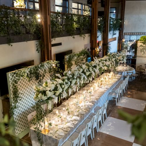 A long dining table setup in a cozy wooden hall with chairs, tea lights, and delicate floral runners, ready for a feast.