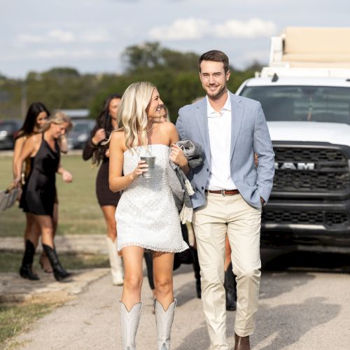 A couple walking with a child and two adults in the back, on a sunny day near a path by the water, smiling and dressed casually.