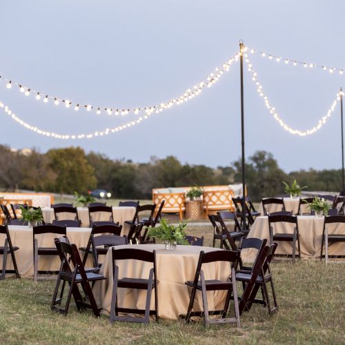 A beachside outdoor wedding reception setup with round tables, chairs, and a canopy/tent in the background, under sunny skies.