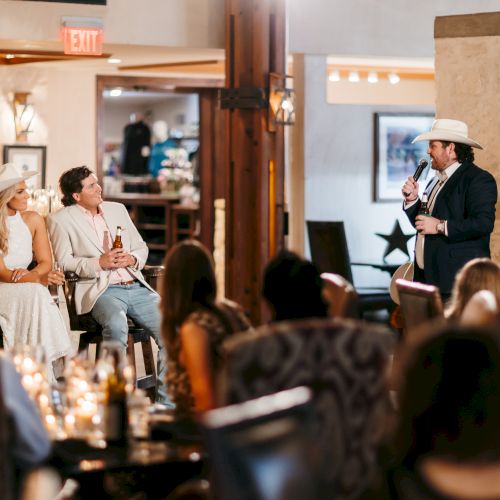 A group of people sits and chats in a cozy caf&eacute;, with warm lighting, plants, and a counter, while a barista serves drinks in the background.