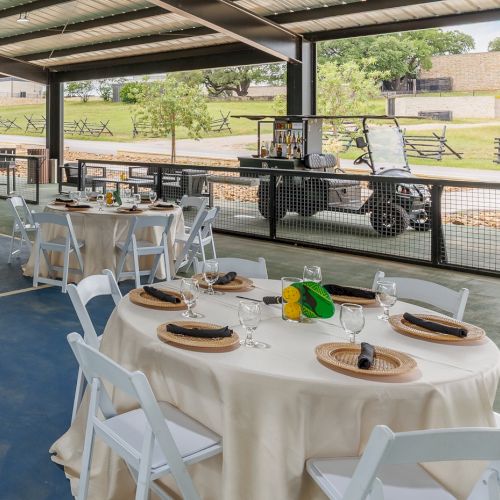 A sunlit kitchen table set for an outdoor meal by a pool, with plates, glassware, wine, and fresh greenery under a shaded patio.