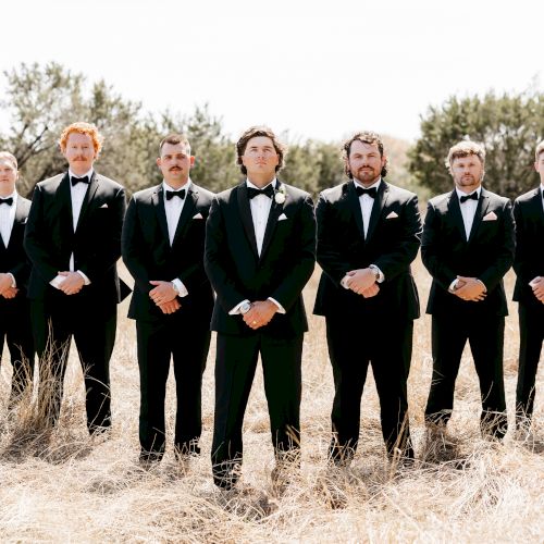 A line of people in dark outfits stands in a field, posing for a group photo, with trees in the background, sunny day.