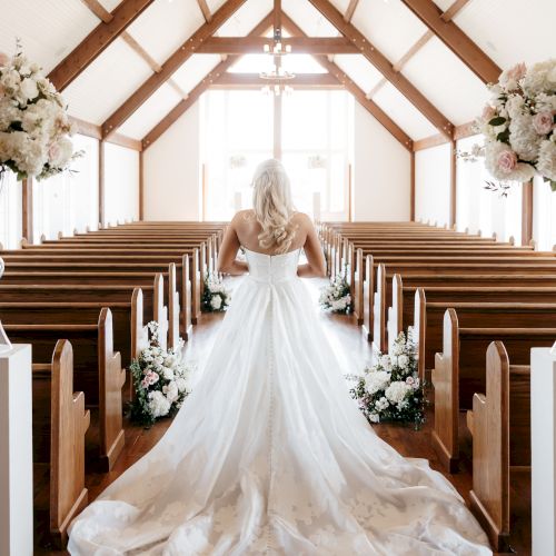 A bride in a long white wedding gown stands at the end of a church aisle, with a carved wooden ceiling and pews on both sides.