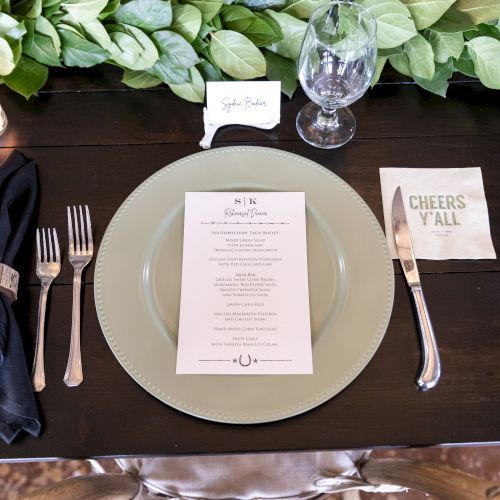 A formal table setting with a dark placemat, a white plate, silverware, a menu card, a name card, and a small bouquet of greenery.