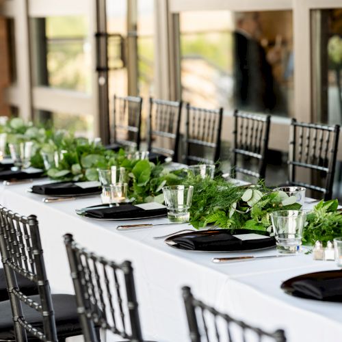 A long outdoor dining area with white chairs and tables along a sidewalk, greenery and lights, a cozy cafe vibe.