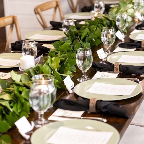A long dining table set for a formal meal with white plates, silverware, glassware, greenery centerpieces, and wooden chairs, in a bright room.