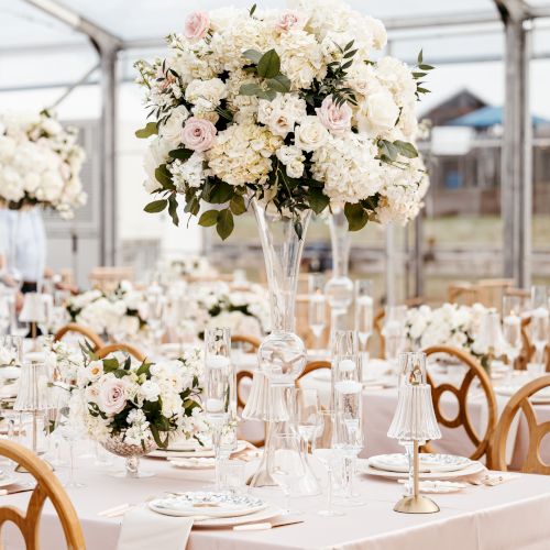 A wedding reception setup with elegant tables dressed in white linens, floral centerpieces, and champagne glasses under a sunny venue.