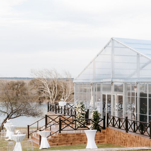A couple gets married outdoors on a sunny day, with guests seated and a glass conservatory in the background.
