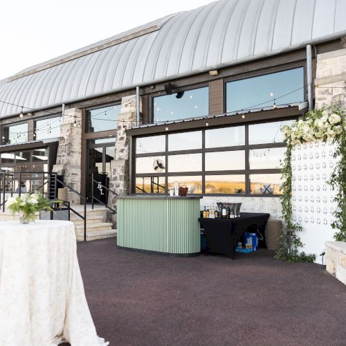 Outdoor event setup with long tables, white linens, and a tent canopy. A snack or beverage station is visible; string lights above add charm.