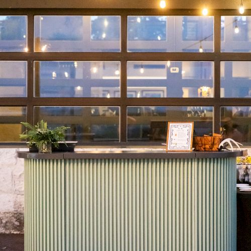 A modern hotel check-in desk with a striped front, plants, and warm lighting, viewed from a reception area by large windows at dusk.