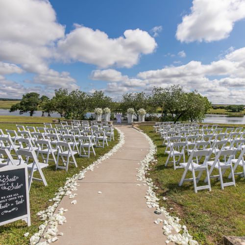 An outdoor wedding setup with white chairs arranged on both sides of a central aisle, petals on the path, a chalkboard sign, and a sunny, cloudy sky.