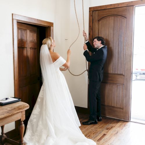 A bride and groom prepare for a ceremony, pulling a long rope from a downed ceiling box, standing in a doorway with wooden floors.