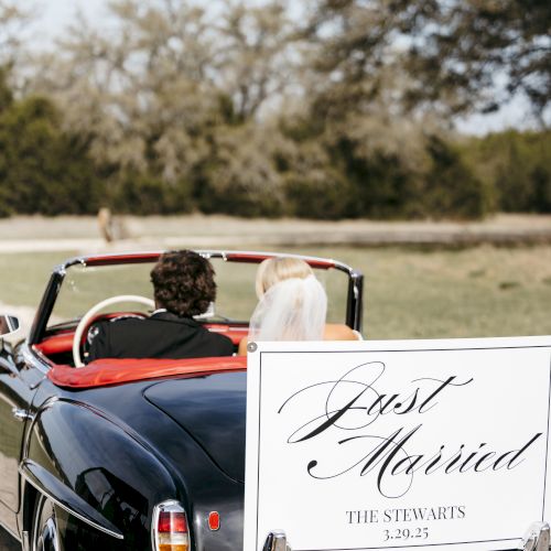 A newlywed couple rides in a vintage black convertible with a white &ldquo;Just Married&rdquo; sign on the back, driving away on a sunny day.