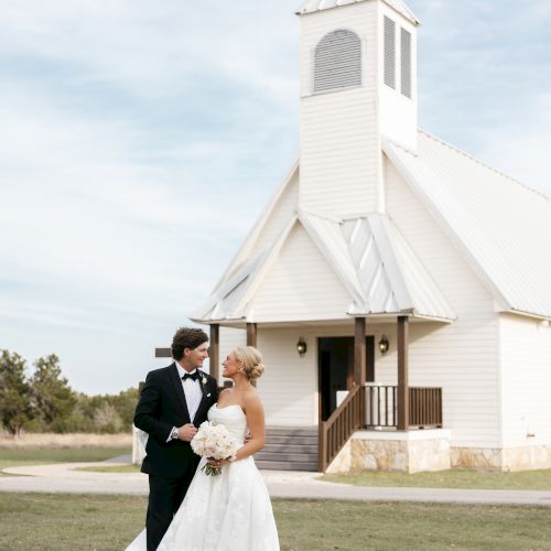 A bride in a white wedding gown and a groom in a black tuxedo stand outside a small white church on a grassy yard, posing for a photo.