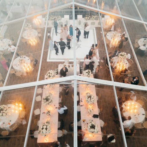 Aerial view of a glass-walled venue with long dining tables, guests mingling, candlelit decor, and a dance floor at the far end, festive ambiance.