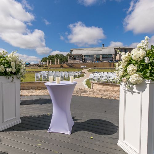 Wedding setup on a wooden deck with two white pedestals holding lush white floral arrangements and a small white-cloth-covered cocktail table, sunny day.