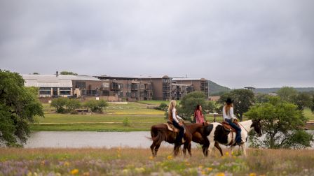Horse riders loop along a riverside with a modern building complex in the background, framed by grassy fields and a cloudy sky.