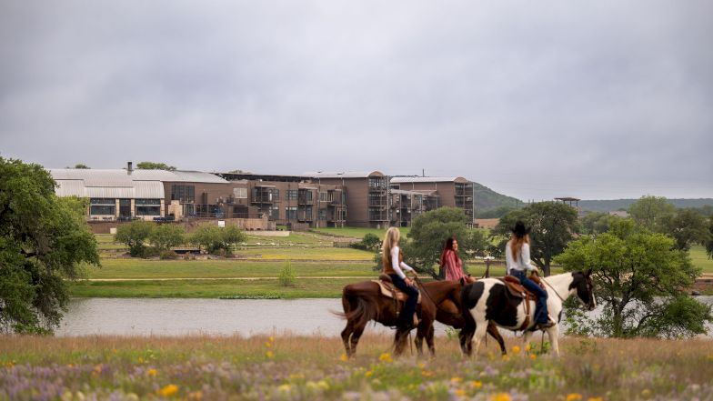 Horse riders loop along a riverside with a modern building complex in the background, framed by grassy fields and a cloudy sky.