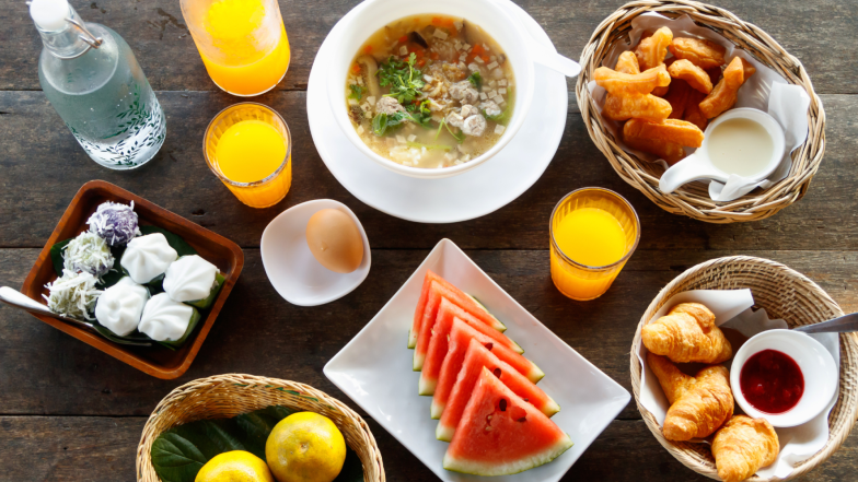A spread of breakfast/brunch: bowls of soup and pastries, watermelon slices, fresh fruit, dumplings, croissants with jam, and fruit juice on a wooden table.