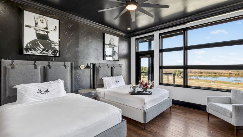 A modern hotel room with two queens, gray upholstered headboards, black-and-white artwork, a large window view, wooden floors, and a seating area.