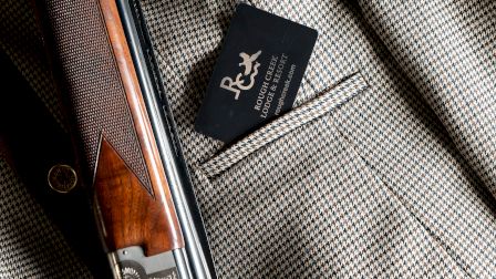 A shotgun rests on a textured coat with a small black card tucked in a pocket, blending vintage leather and metal details, stylishly arranged.