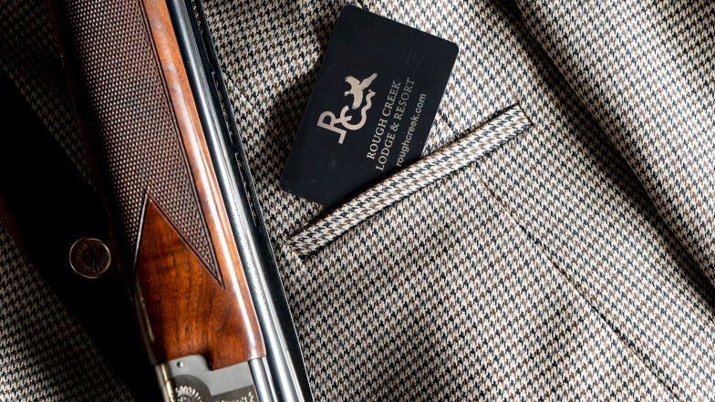 A shotgun rests on a textured coat with a small black card tucked in a pocket, blending vintage leather and metal details, stylishly arranged.