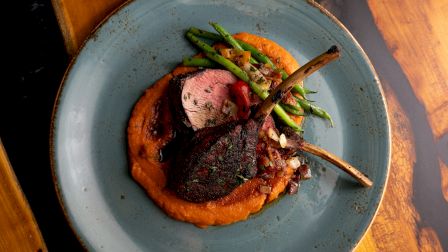 A plated grilled meat chop with char, sits on a orange pur&eacute;e, garnished with green beans on a blue plate, cozy dinner vibe.