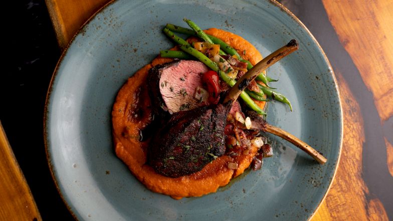 A plated grilled meat chop with char, sits on a orange pur&eacute;e, garnished with green beans on a blue plate, cozy dinner vibe.