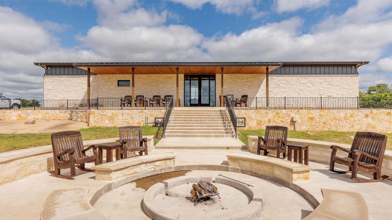 A modern single-story building with a wide entrance, stone walls, and a central fire pit circle surrounded by wooden chairs on a concrete patio. End.