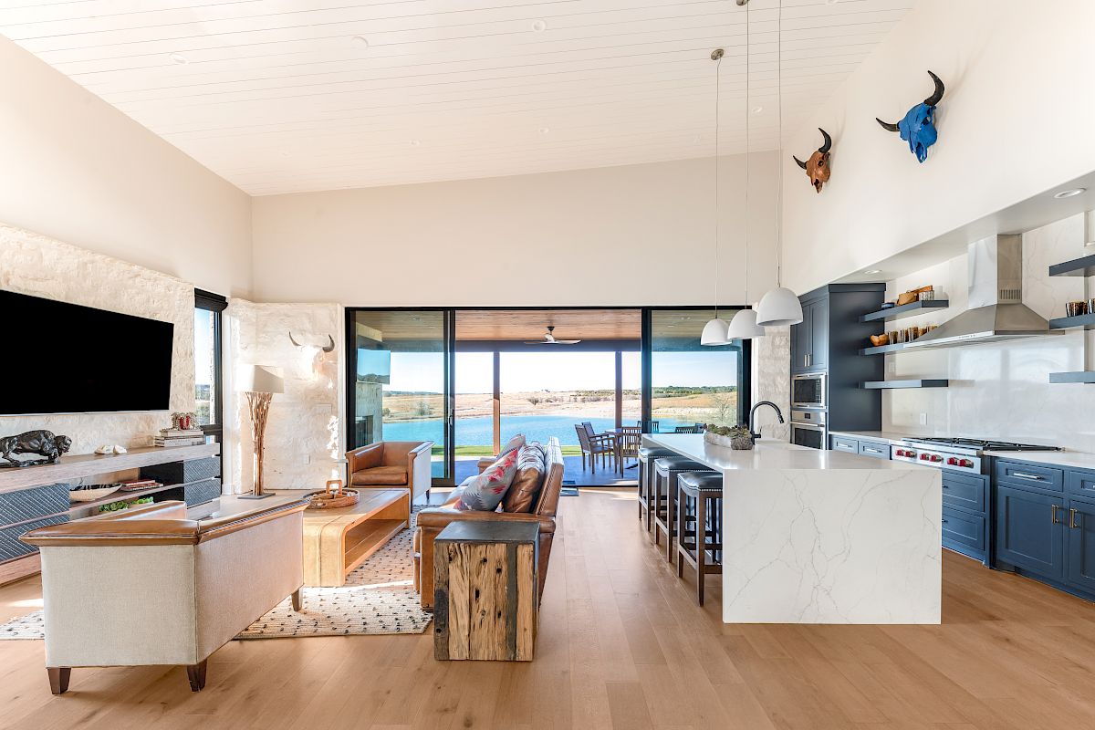 Spacious, modern open-plan living room and kitchen with a big island, blue cabinets, wood floors, and a view of the sea through sliding doors. Ends with a period.
