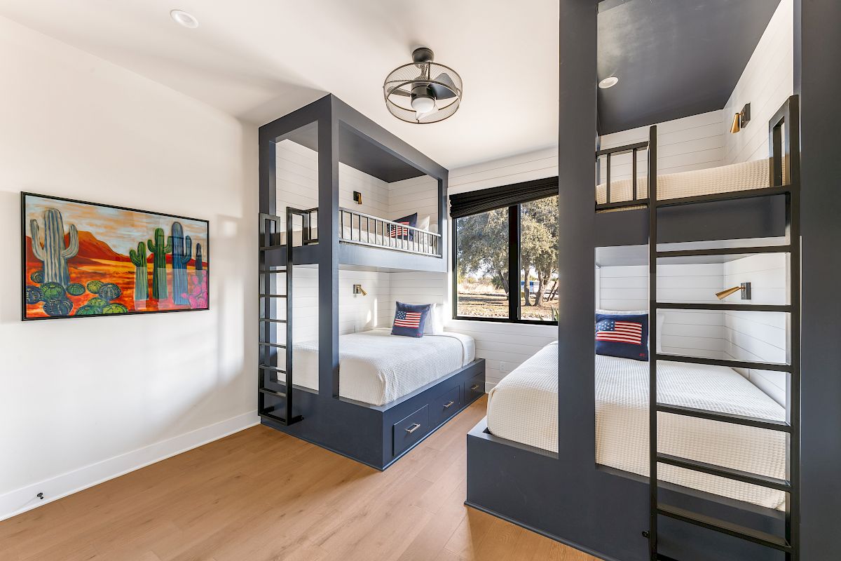 Two bunk beds with built-in stairs and drawers, a bright modern bedroom with a large window, colorful art, and light wood flooring.