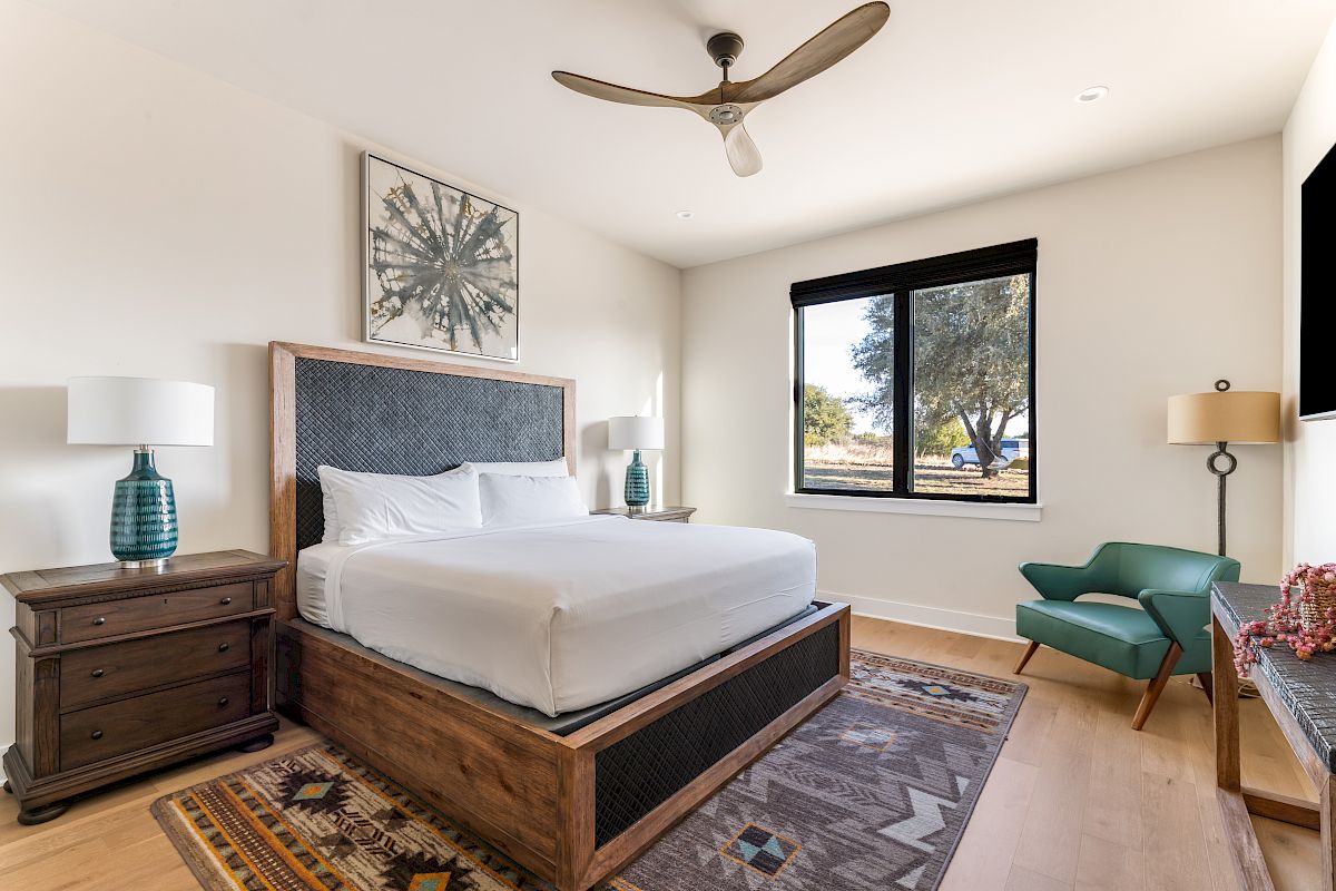 A bright, modern bedroom with a wooden bed, white linens, two nightstands, lamps, a ceiling fan, a window view, a green chair, and a patterned rug, stylishly cozy.