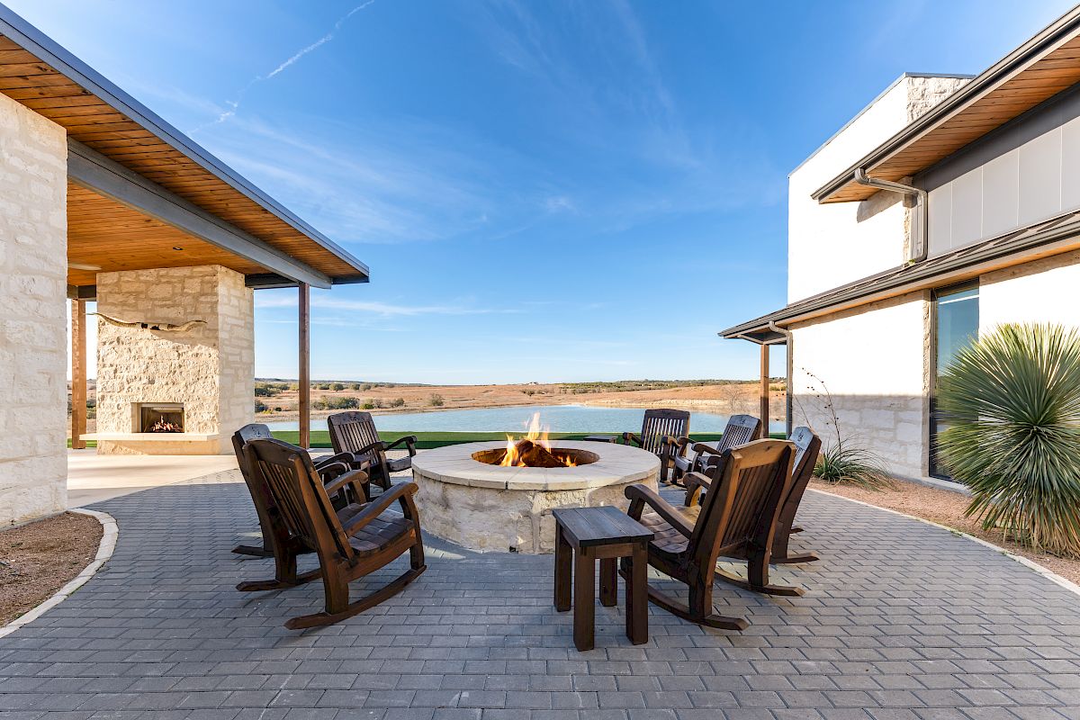 Outdoor modern courtyard with stone buildings, a circular fire pit, and wooden chairs surrounding it, overlooking a calm body of water under a clear sky.