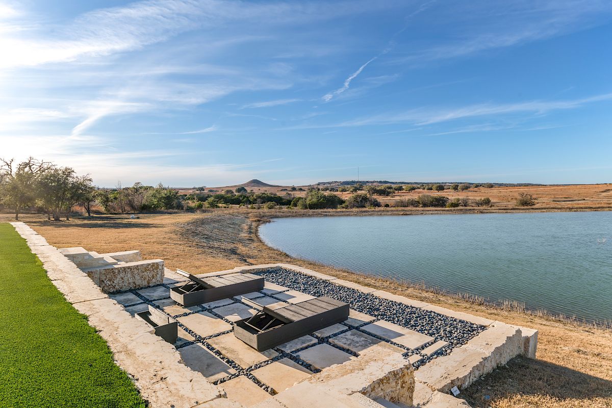 A lakeside patio with modern stone seating and gravel edging, green lawn on the left, calm blue water, and a clear sky.