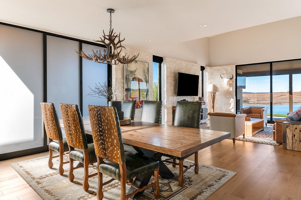 A modern dining room with a wooden table, eight chairs, antler chandelier, large glass walls, and a scenic outdoor view ending with a sentence.