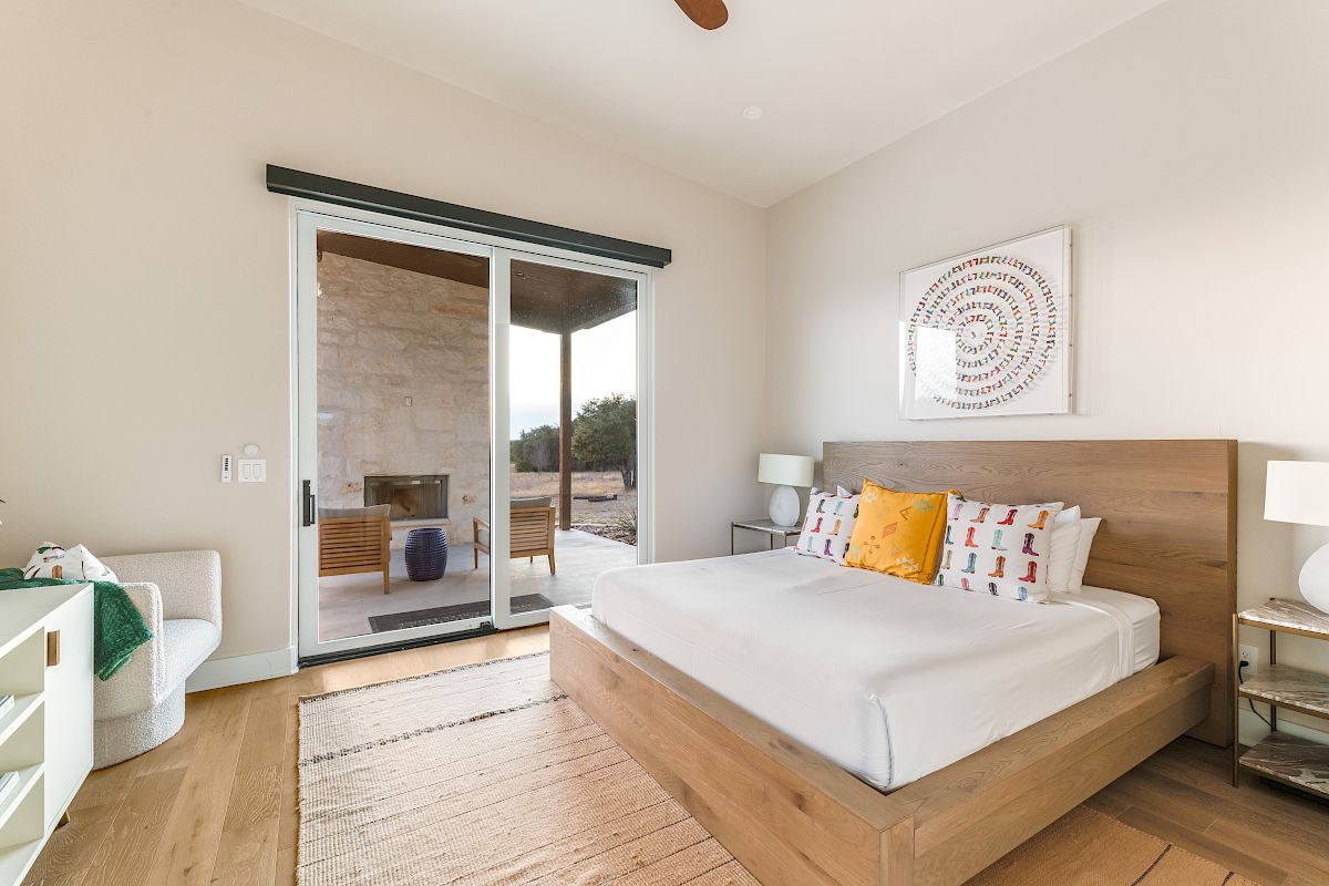 A modern bedroom with a wooden bed, white bedding, decorative pillows, nightstands, a sliding glass door to an outdoor patio, and warm natural light.