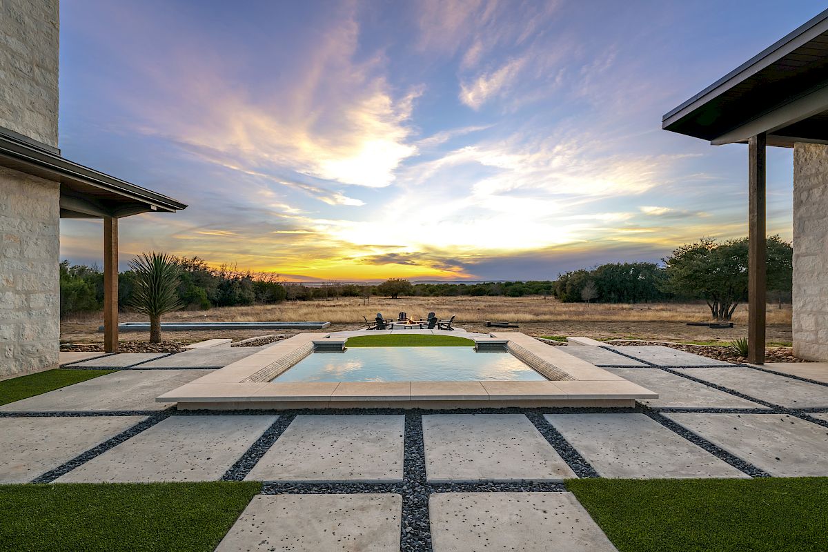 A luxurious modern courtyard with a rectangular pool, stepping stone paths, manicured lawn, and a sunset sky between two stone buildings.