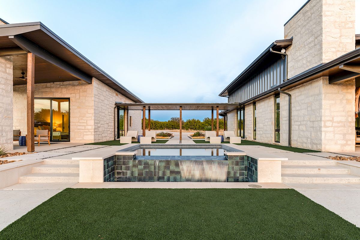 Luxurious modern home with a central rectangular pool, stone walls, expansive glass doors, and a manicured lawn leading to an open-air seating area.
