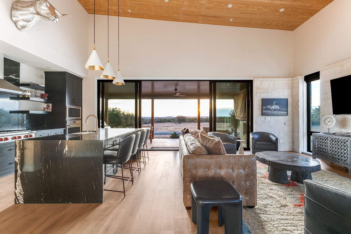 Luxurious modern open-plan living with a polished kitchen, island seating, and panoramic ocean views through sliding glass doors at sunset.