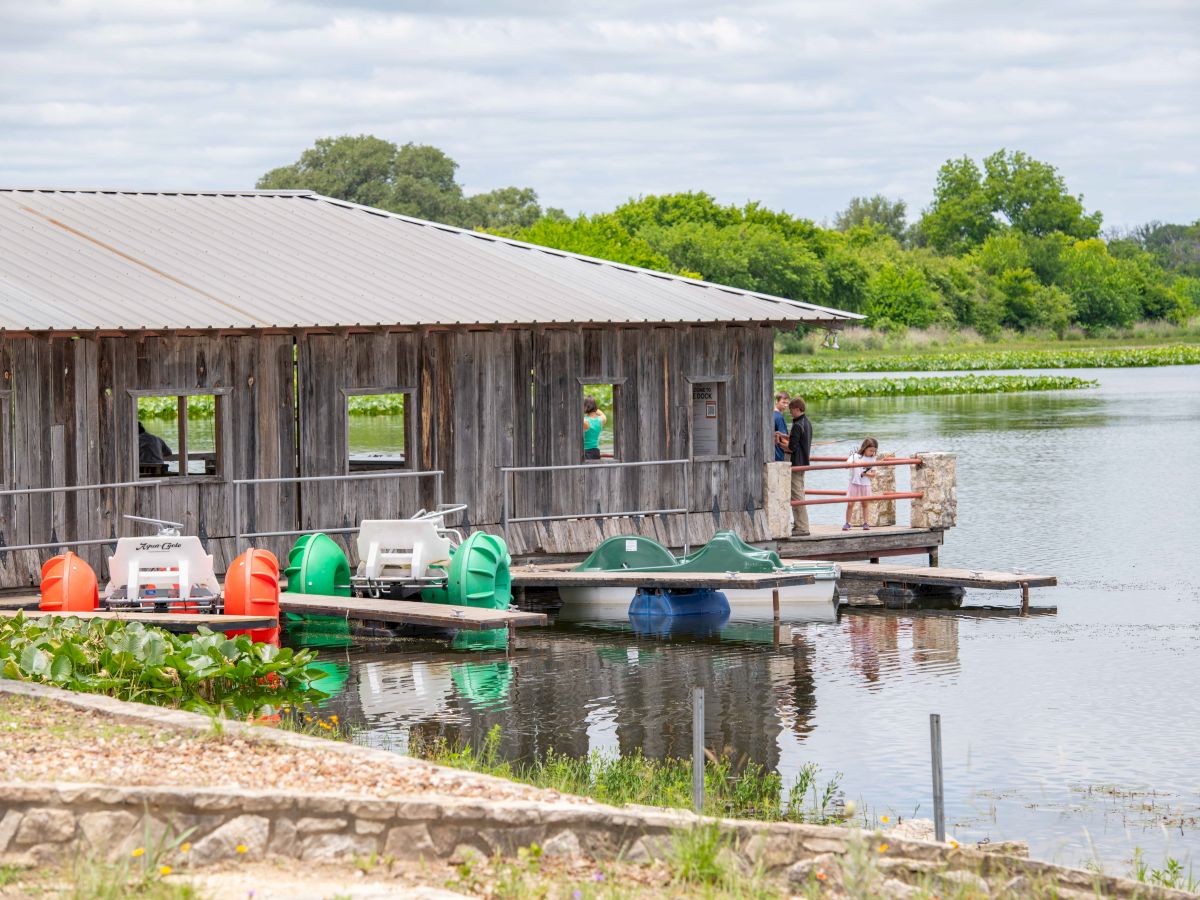 A rustic wooden boathouse on the water with colorful gondola-like boats and a person standing on a dock, calm scenery and green trees nearby.