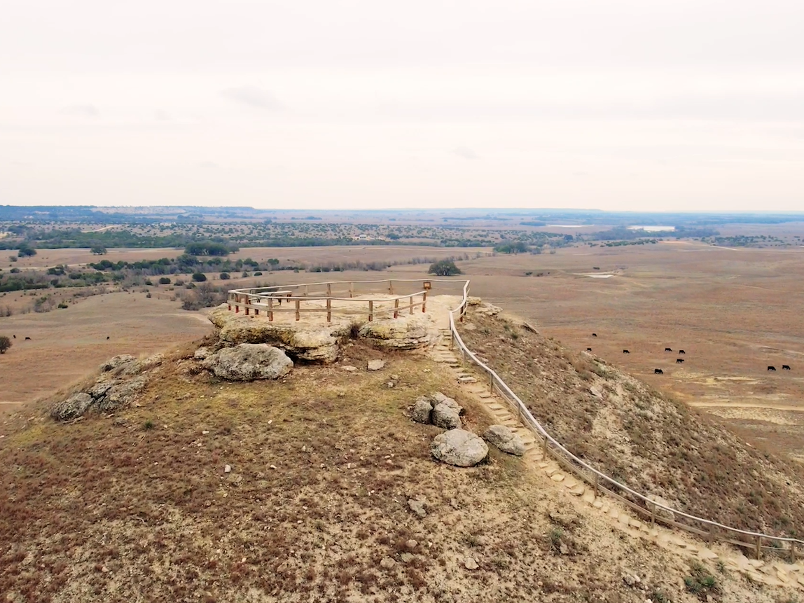 A hilltop viewpoint with a wooden walkway and fence, overlooking a wide, dry savanna landscape stretching to the horizon.