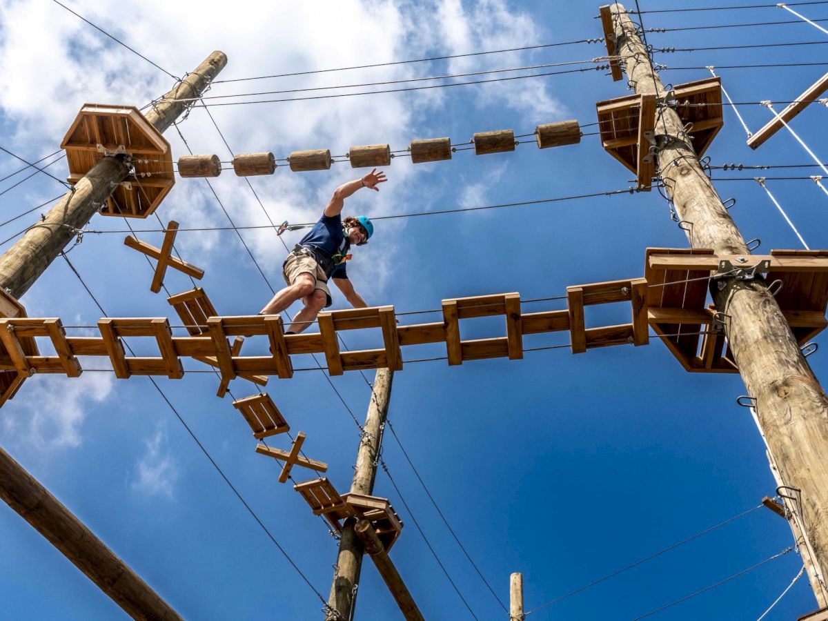 A person crosses a high wooden obstacle course built on tall poles, suspended bridges and ropes against a bright blue sky.