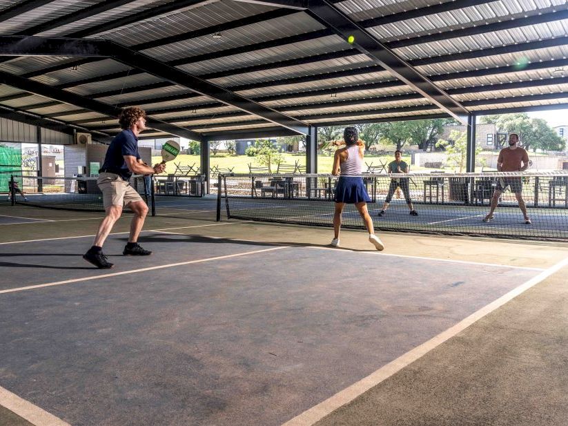 People playing a game on a covered outdoor court, with nets and a ball in motion, as others watch from the sidelines, all focused and active.